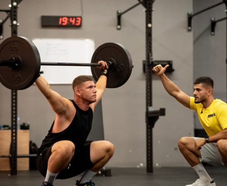 Athlete performing weightlifting exercise under trainer's guidance in a gym setting.